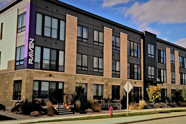 A modern three-story building in Madison, WI, with large windows, labeled "Raven" on a vertical sign. The facade features a mix of light and dark tones, complemented by recent exterior renovations with stone and brick accents. Landscaping includes shrubs, grasses, and a path. A fire hydrant is visible on the sidewalk.