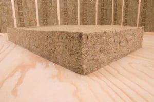 A thick rectangular slab of insulation material (likely mineral wool or rock wool) is displayed on a light-colored wooden surface. In the background, vertical wooden studs and additional insulation material can be seen lining the walls—perfect for exterior renovations in Madison, WI.