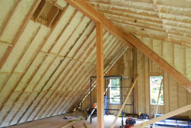 An unfinished attic with wooden beams and walls insulated with spray foam. The space has a small window and construction tools scattered on the floor, including a ladder, a saw, and a toolbox. This project in Madison, WI is part of broader exterior renovations.