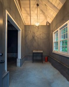 A room with a high, angled ceiling and muted blue-grey walls. There is a spherical chandelier hanging from the ceiling. The room features two windows on one wall and an open doorway leading to another space. A small table and red bucket are under the windows, hinting at recent exterior renovations in Madison, WI.