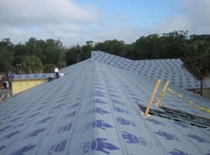A large roof in Madison, WI, covered with blue synthetic underlayment sheets labeled 'REX,' awaits shingling. Wooden planks and tools are visible, surrounded by trees under a cloudy sky, showcasing the exterior renovations in progress.
