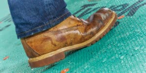 Close-up shot of a person's foot wearing a well-worn brown leather shoe. The shoe appears scuffed and damp, and the person is standing on a green surface, possibly related to exterior renovations in Madison, WI, with red markings or buttons. The person is wearing dark blue jeans.