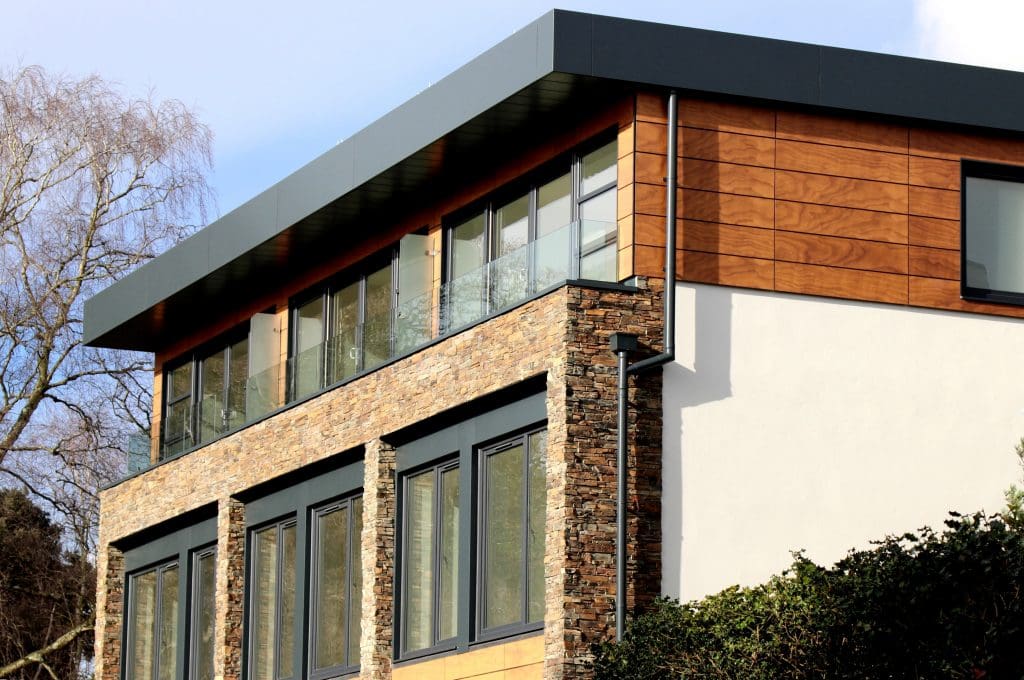 A modern, multi-story house in Madison, WI featuring a mix of materials. The upper floor has large windows and wood siding, while the lower level boasts stone walls and smaller windows. Recently updated with exterior renovations, the house includes a sleek, flat roof and is surrounded by trees and bushes.