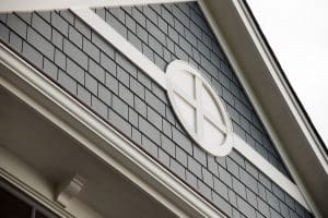 Close-up of a house exterior in Madison, WI, featuring dark gray shingle siding and white trim. There is a round, white, four-pane window near the peak of the roof. The shingles are neatly arranged, creating a clean and stylish look following recent exterior renovations. The roofline is partially visible.
