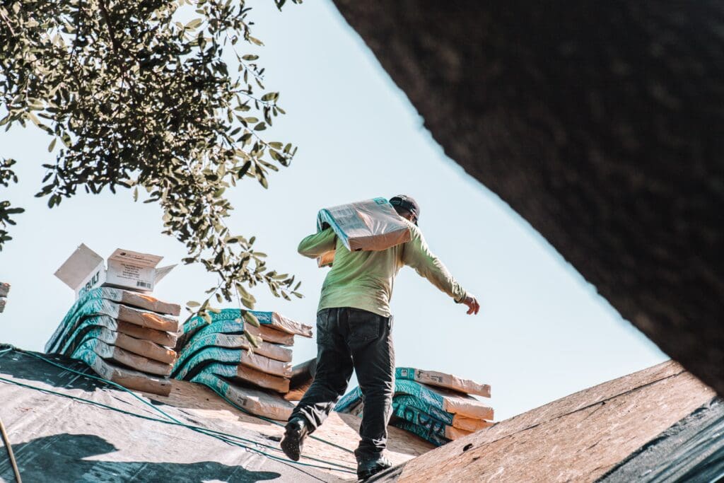 A construction worker in a green shirt and dark pants carries a bag of roofing material on his shoulder while walking on a slanted roof. Several stacks of similar bags are placed beside him, hinting at ongoing exterior renovations. Tree branches frame the scene under a clear Madison, WI sky.