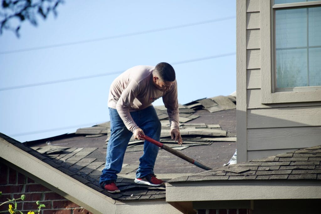 A person wearing a long-sleeve shirt, jeans, and sneakers is standing on a house roof in Madison, WI, using a tool to remove shingles. Part of the roof appears damaged. The siding and windows of the house are visible on the right side of the image, indicating ongoing exterior renovations.