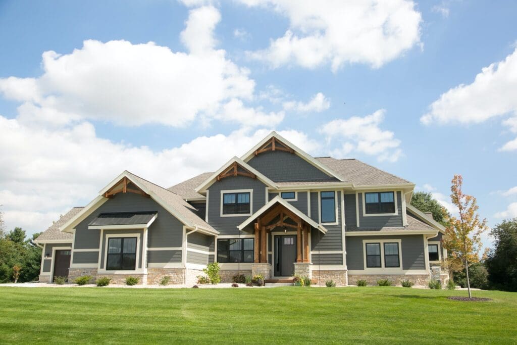 A large, modern two-story house in Madison, WI with a gray exterior, stone accents, and multiple gable roofs. The house features a prominent entrance, large windows, and a well-manicured lawn with a few small trees. The sky is clear with scattered clouds—perfect for showcasing elegant exterior renovations.