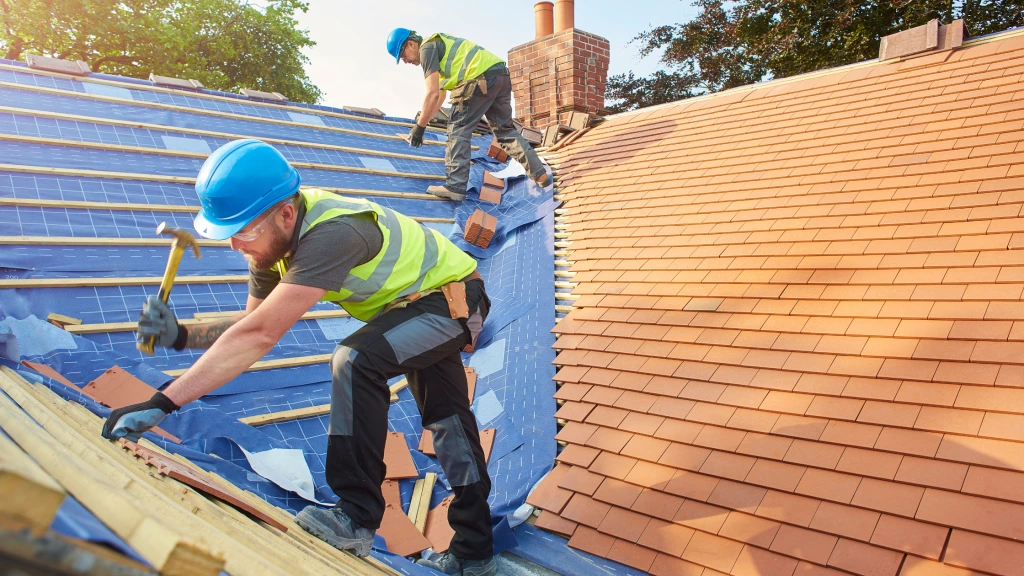 Two construction workers in safety gear install roof tiles on a house, showcasing the roofing benefits of expert roofing services. One worker hammers tiles in place while the other arranges tiles up the roof under sunlight and trees.