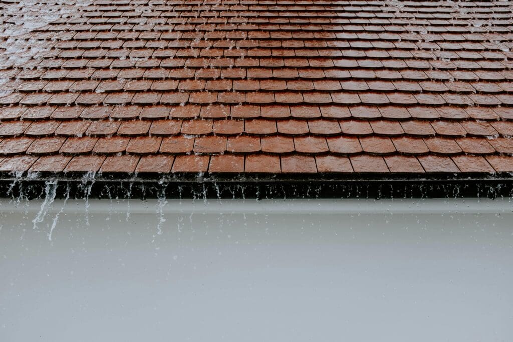 Rainwater cascades off the edge of a sloped, red-tiled roof, highlighting the importance of a well-maintained homes roof. The neatly arranged tiles send droplets streaming over the edge, creating a dynamic scene against the gray sky, underscoring why upgrading is essential for enduring beauty and protection.