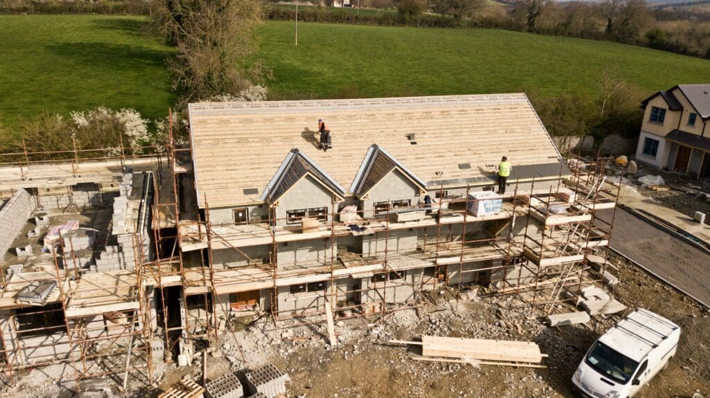 Aerial view of a two-story house under construction. Workers are upgrading the homes roof, securing materials with precision. Scaffolding surrounds the building, and construction debris is scattered below. A green field and additional houses form the backdrop, highlighting the importance of progress.