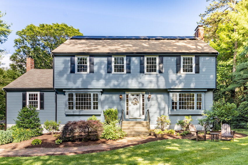 The two-story blue home boasts a symmetrical front, large windows, and a central entrance. Wrapped in fiber cement siding, its surrounded by greenery and trees, featuring a manicured lawn with two wooden chairs in the yard under a clear blue sky.
