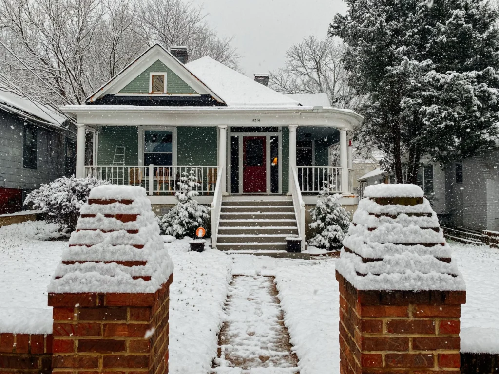 A cozy, green house with a white roof and front porch in Madison, WI is surrounded by snow. Snow-covered stairs lead up to the porch, and a path flanked by two brick pillars extends to the sidewalk. Recent exterior renovations complement the snow-blanketed trees and neighboring houses, creating a serene, wintry scene.