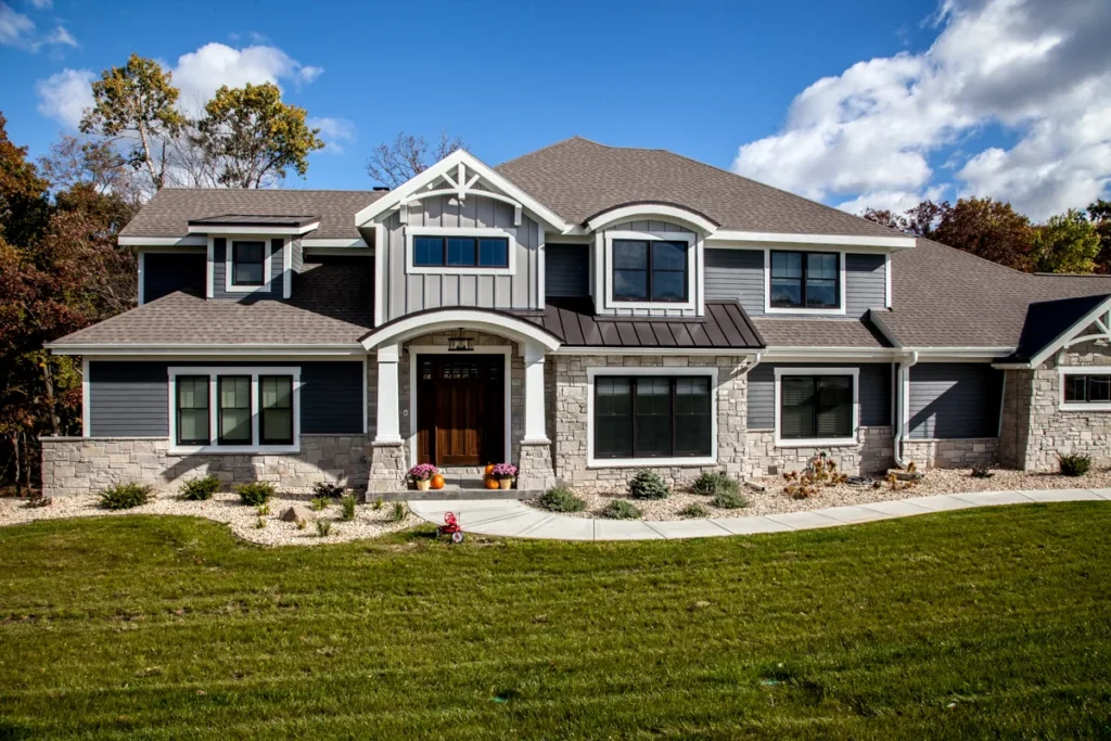 A large, modern two-story house in Madison, WI features a gray exterior with stone accents and white trim. The entryway boasts a dark wooden door with sidelights. Recent exterior renovations include a landscaped front yard with a stone path, green grass, and flower pots under a partly cloudy sky.
