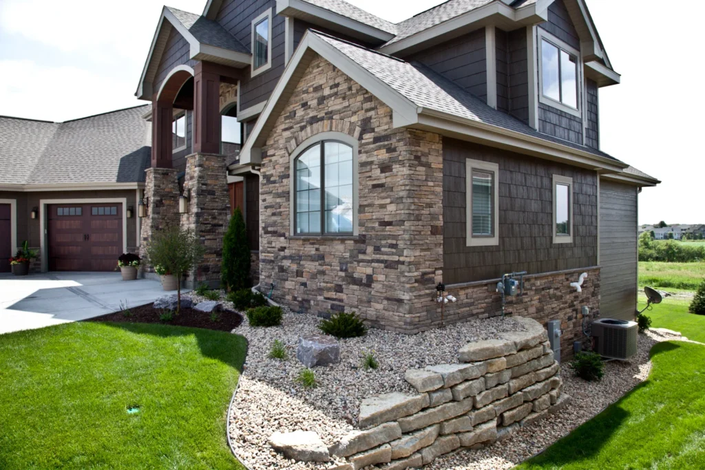 A modern house in Madison, WI with brown siding and stone accents, featuring large windows and a well-maintained lawn. Recently completed exterior renovations include a small stone retaining wall with shrubbery and a neatly landscaped front yard with a concrete driveway leading to a three-car garage.
