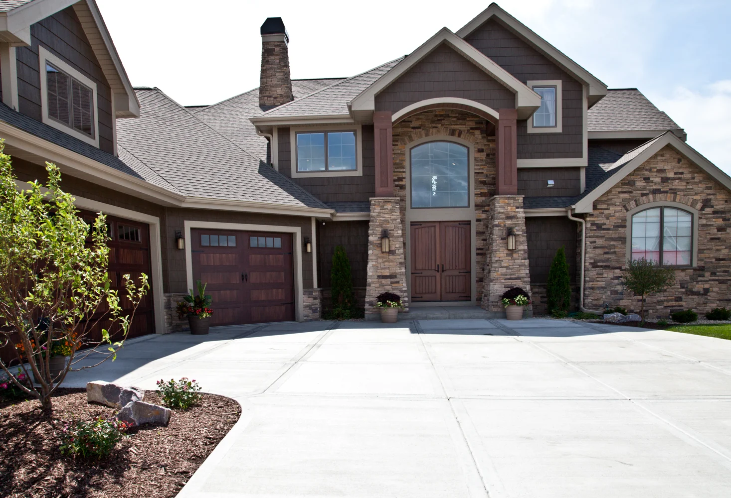 A large two-story house with brown siding and a stone facade, featuring a two-car garage with wooden doors, two wooden front doors with glass panels above them, and neatly landscaped surroundings including a tree and flower pots. Recently completed exterior renovations have made this Madison, WI home even more inviting.