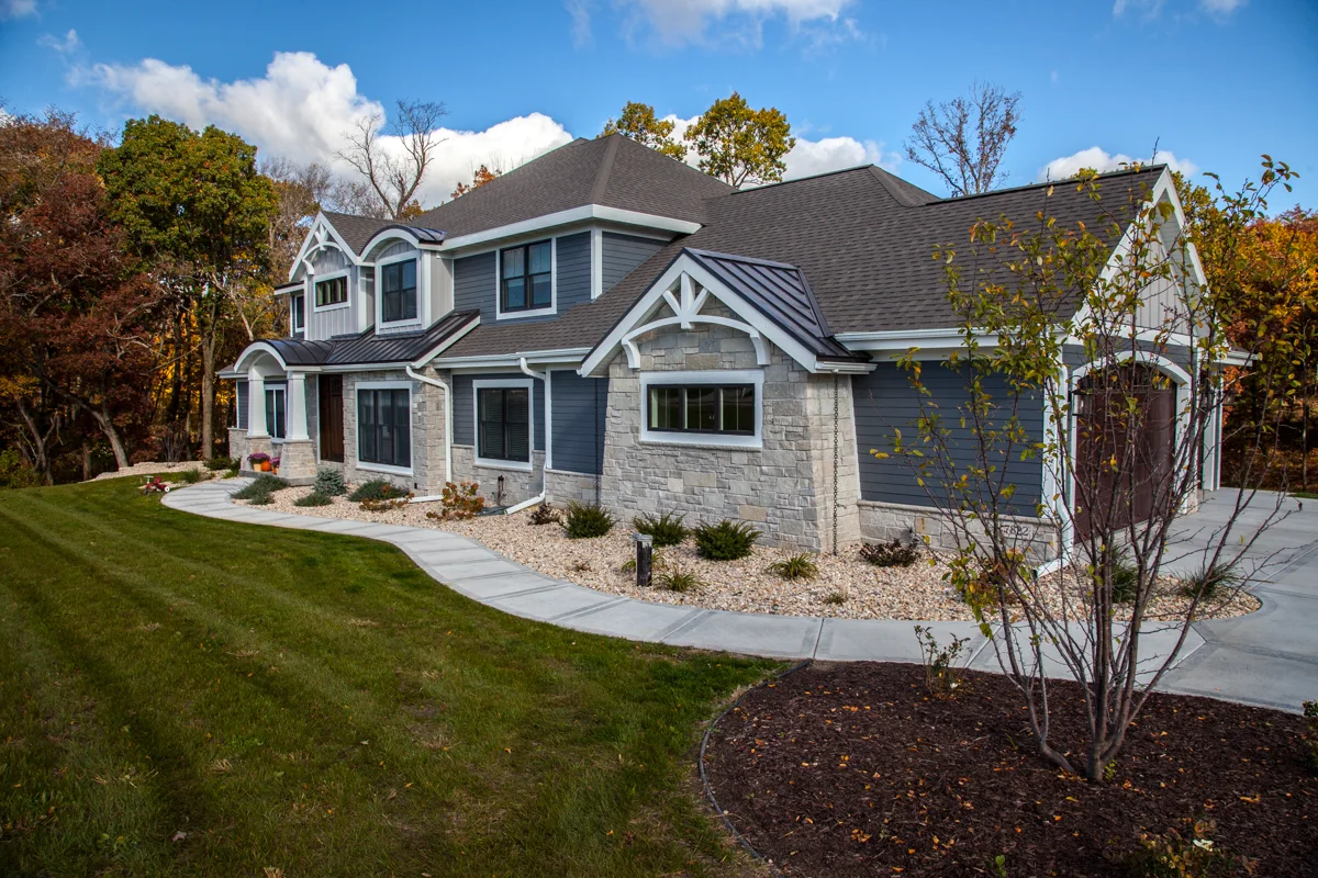 A modern two-story house with gray siding, stone accents, and a black shingled roof in Madison, WI. The home features a manicured lawn, a curved walkway, and surrounding trees with autumn foliage. A three-car garage is on the right side, showcasing recent exterior renovations under partly cloudy skies.