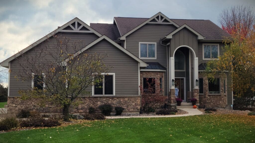 A modern two-story house with gray siding, stone accents, and a covered entrance stands proudly in the heart of Madison, WI. The yard features a well-maintained lawn, a pathway leading to the front door, and a few trees with autumn foliage. Recent exterior renovations complete the polished look under the cloudy sky.