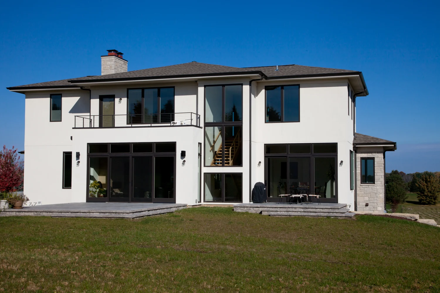 A modern two-story house in Madison, WI, with a renovated white exterior, large glass windows, and a spacious backyard with green grass. The house features a balcony, a stone chimney, and a patio area with outdoor furniture, set against a clear blue sky.