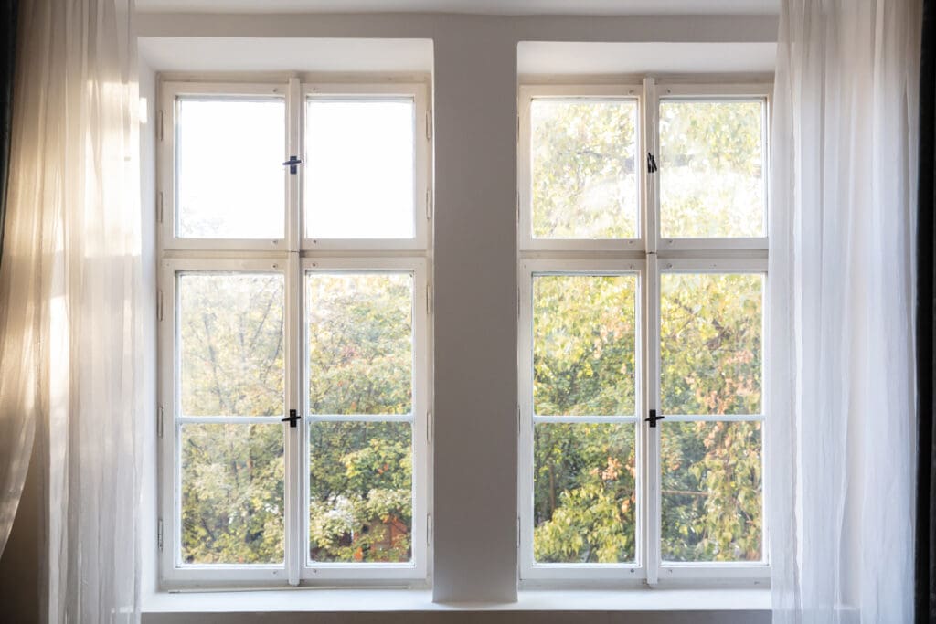 Two tall, energy-efficient windows with white frames and multiple panes let in soft natural light. Sheer white curtains frame the windows, while green, leafy trees outside benefit from excellent window insulation.