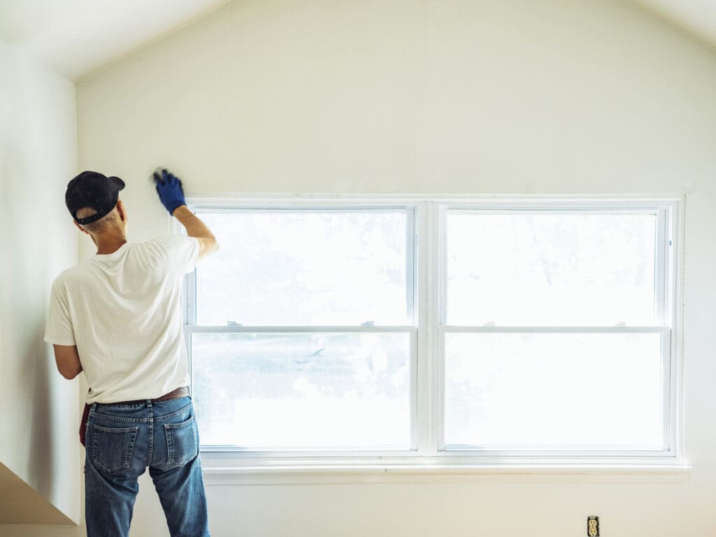 A person wearing a white shirt, jeans, and a black cap is cleaning or preparing a white wall above energy-efficient windows in a bright room, using a gloved hand.