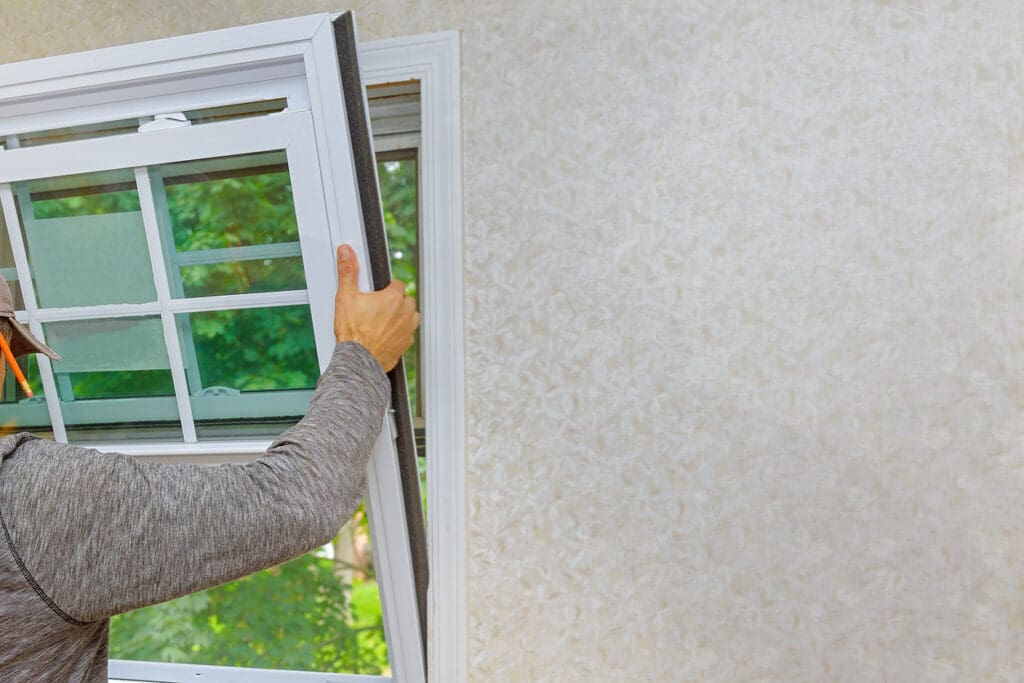A person installs or removes a white-framed, grid-paned energy-efficient window. Only their arm and part of their torso are visible. The window overlooks a green, leafy outdoor area.
