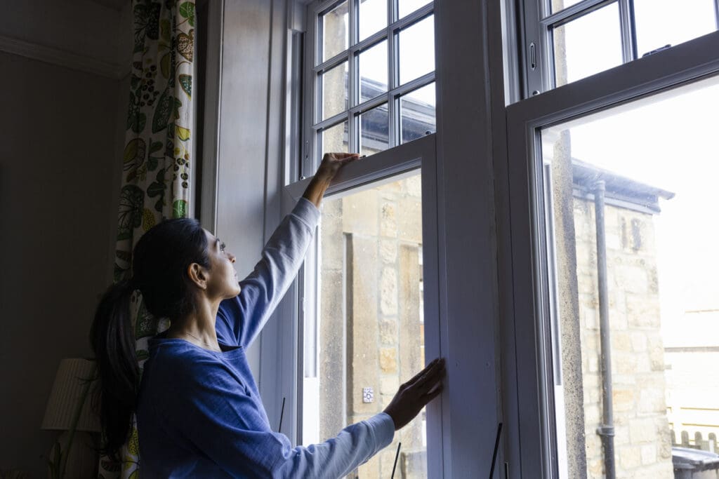A woman with dark hair in a ponytail, wearing a blue long-sleeve shirt, opens an energy-efficient window in a bright room, letting in fresh air and natural light. The view outside reveals a stone building.
