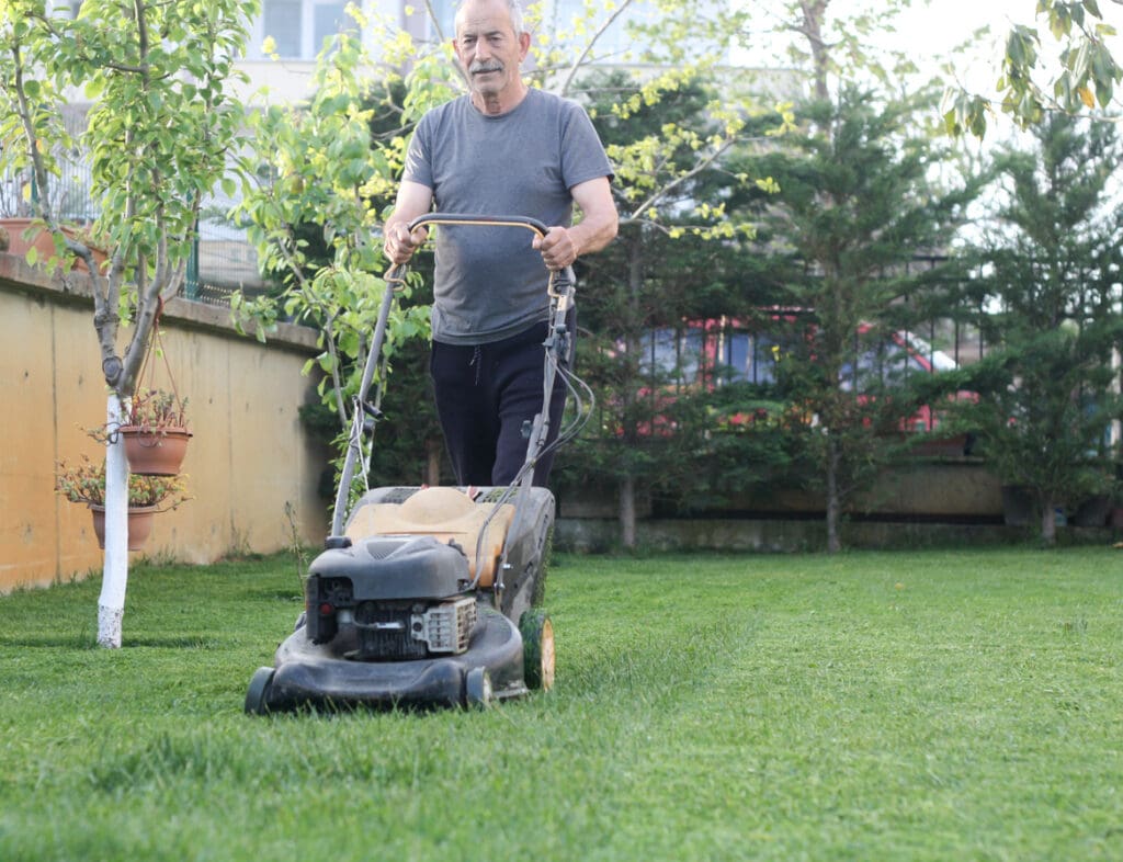 An older man in a grey t-shirt and black pants is mowing the lawn in a backyard garden with green grass, trees, and a yellow wall—enhancing the curb appeal as part of simple exterior home improvements.