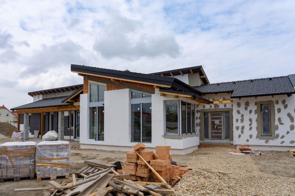 A modern house under construction showcases large windows, exposed wooden beams, and partially finished exterior walls—exemplifying impressive curb appeal. Construction materials like bricks and wooden planks are scattered in the foreground.