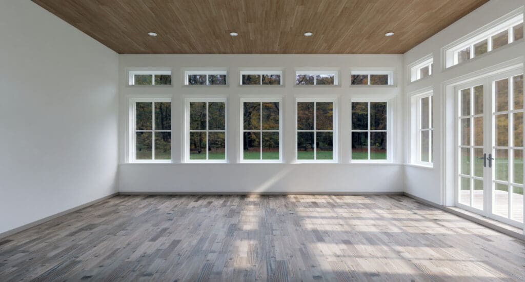 A bright, empty room with large windows, a wooden floor, and a wood-paneled ceiling features energy-efficient windows that offer a clear view of green trees and a grassy yard outside.