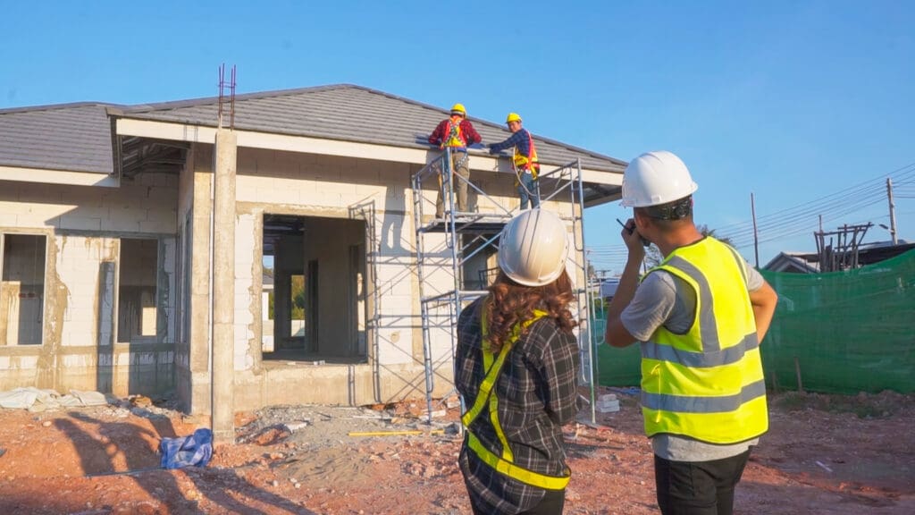 Two construction workers on scaffolding handle exterior home improvements on a house under construction, while two supervisors in safety vests and helmets observe and communicate from the ground.