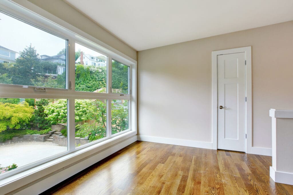 Bright, empty room with large windows featuring energy-efficient windows, wooden floor, beige walls, white trim, and a white door. Outside the windows, green trees and a hillside with houses are visible.