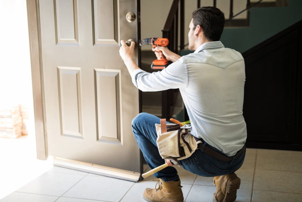 A man kneels while installing or repairing a front door with a power drill, enhancing curb appeal. He wears jeans, boots, and a tool belt in a bright entryway near stairs, working on exterior home improvements.