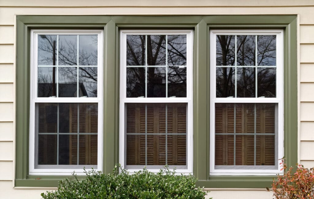 Three vertical, energy-efficient windows with green trim set in a cream-colored wall reflect bare tree branches. Brown wooden blinds are partially visible behind the glass, with green shrubs in front of the window insulation.