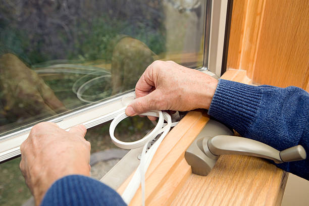Person in a blue sweater applying weatherstripping tape to the inside edge of a wooden window frame, improving window insulation and preparing for more energy-efficient windows.
