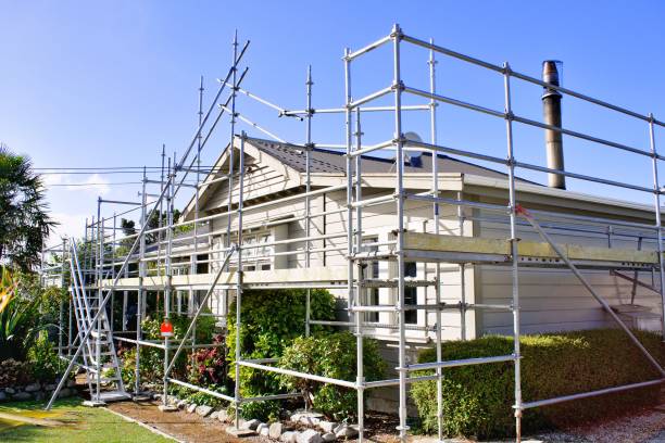 A beige house surrounded by metal scaffolding, indicating exterior renovations to increase value, with a blue sky in the background and plants along the front yard.