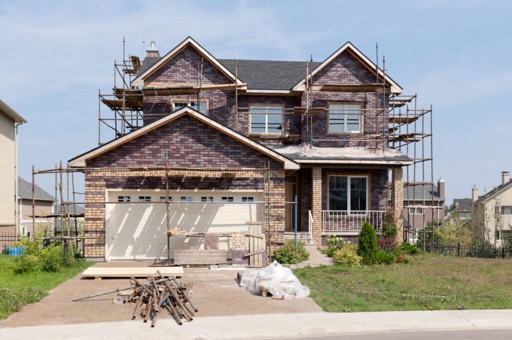 A two-story house undergoing a Whole-House Exterior Makeover, with scaffolding surrounding the home exterior, building materials and tools in the driveway, and a front yard featuring grass and bushes.