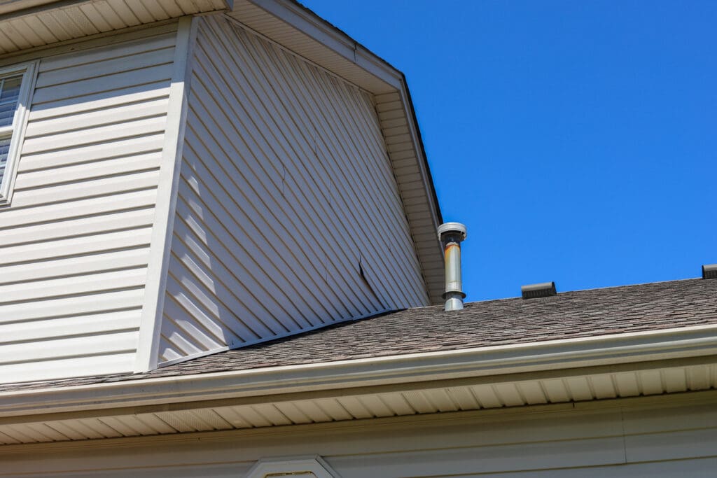 Close-up view of a house’s exterior, showing light-colored siding, a sloped shingled roof, and a metal vent pipe under a clear blue sky—ideal for assessing when to replace siding or plan siding repair. Part of a window and roof eaves are also visible.