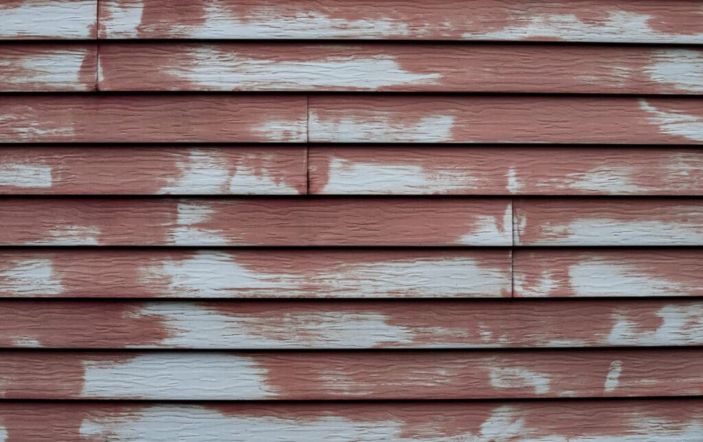 Close-up of weathered horizontal wooden siding with peeling red paint, revealing patches of bare wood and white primer—signs that siding repair or even siding replacement may soon be needed. The surface appears rough and aged.
