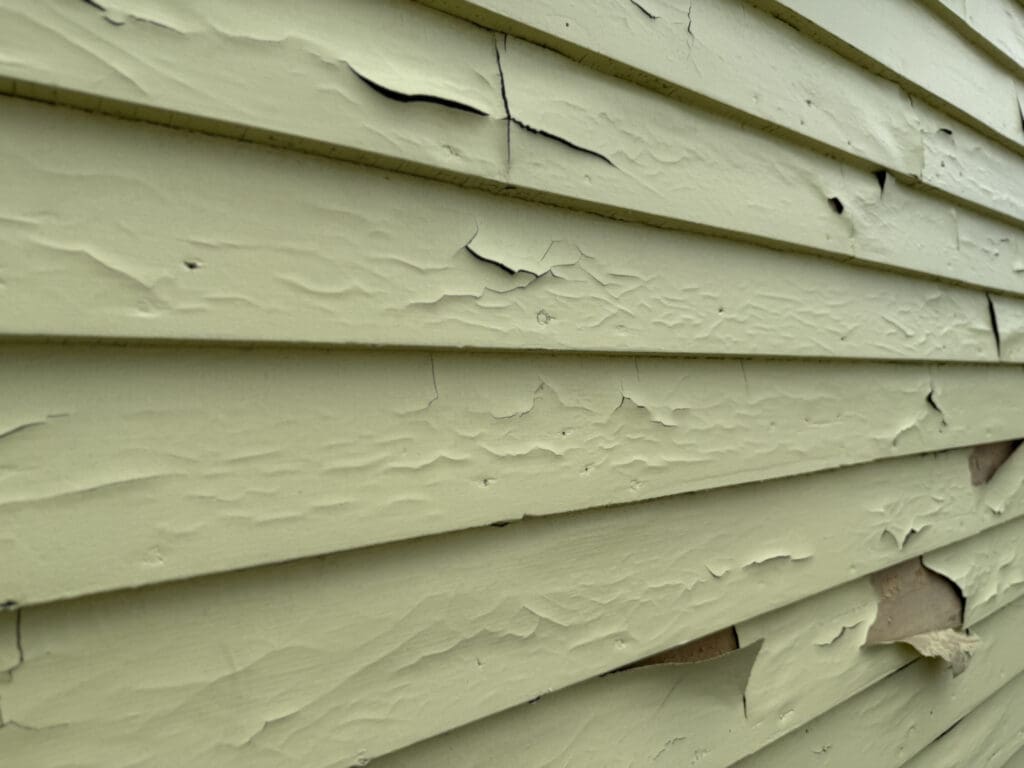 Close-up of light green wooden siding with peeling, cracked paint and visible signs of weathering—clear indications that siding repair or even siding replacement may be needed.