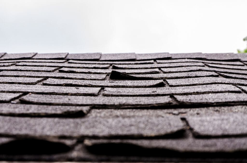 Close-up view of a dark asphalt shingle roof with several shingles lifting or curling upward—clear roof warning signs that indicate potential damage or wear. An overcast sky looms in the background, highlighting the need for roof repair.