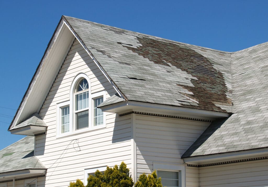 A white house with a gable roof shows warning signs like worn and missing shingles, signaling the need for immediate repair beneath a clear blue sky.