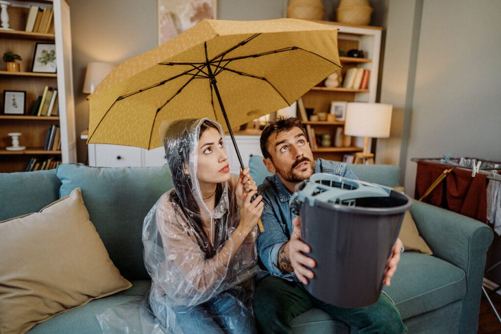 A man and woman sit on a couch in a living room. The woman wears a rain poncho and holds a yellow umbrella, while the man catches water dripping from the ceiling—a clear warning sign that roof repair is needed. Both look up with concerned expressions.
