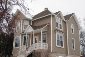 A two-story beige house with white trim and a gabled roof stands proudly in Madison, WI. The exterior renovations include a small porch with a white railing at the entrance. Multiple windows overlook the snowy landscape, with bare trees and a parked car partially visible.