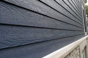 Close-up of a house exterior in Madison, WI, showcasing dark blue horizontal wooden siding. The bottom portion features a beige trim transitioning into a stone base. Various textures and materials are evident, providing visual contrast. Partial view of a window on the right highlights recent exterior renovations.